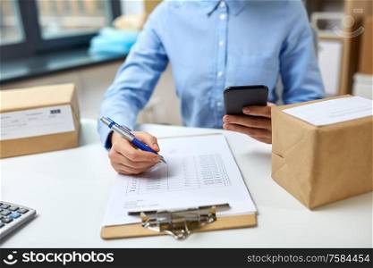 delivery, mail service, people and shipment concept - close up of woman with smartphone and parcel boxes filling checklist on clipboard at post office. woman with smartphone and clipboard at post office