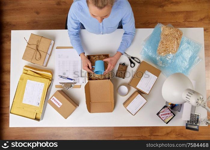 delivery, mail service, people and shipment concept - close up of woman packing mug to parcel box with straw filler at post office. woman packing mug to parcel box at post office