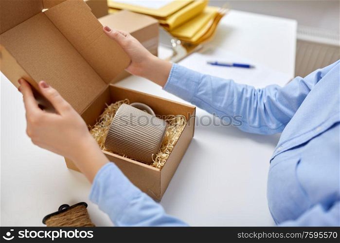 delivery, mail service, people and shipment concept - close up of woman packing mug to parcel box with eco straw filler at post office. woman packing mug to parcel box at post office