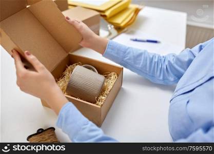delivery, mail service, people and shipment concept - close up of woman packing mug to parcel box with eco straw filler at post office. woman packing mug to parcel box at post office