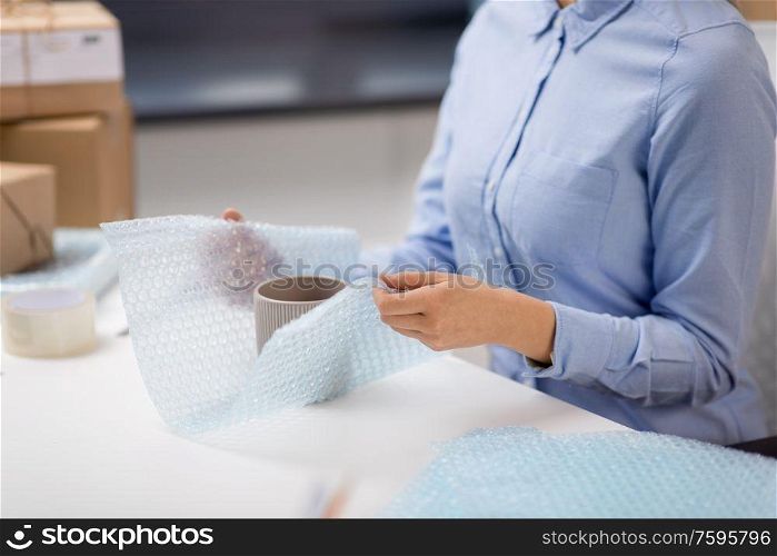 delivery, mail service, people and shipment concept - close up of woman packing mug and it wrapping into protective bubble wrap at post office. woman packing mug to parcel box at post office