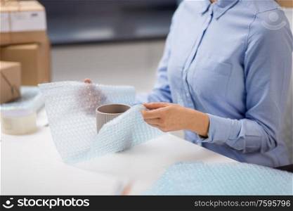 delivery, mail service, people and shipment concept - close up of woman packing mug and it wrapping into protective bubble wrap at post office. woman packing mug to parcel box at post office