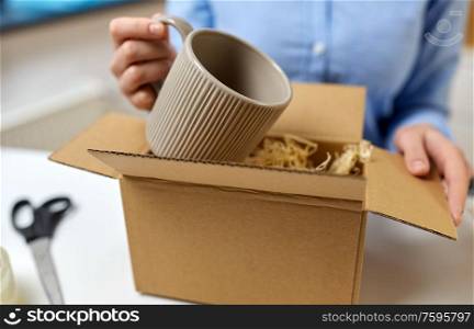delivery, mail service, people and shipment concept - close up of woman packing mug to parcel box with straw filler at post office. woman packing mug to parcel box at post office