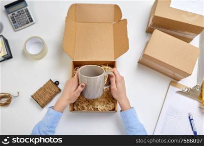 delivery, mail service, people and shipment concept - female hands packing mug to parcel box with straw filler at post office. hands packing mug to parcel box at post office