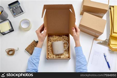 delivery, mail service, people and shipment concept - female hands packing mug to parcel box with straw filler at post office. hands packing mug to parcel box at post office