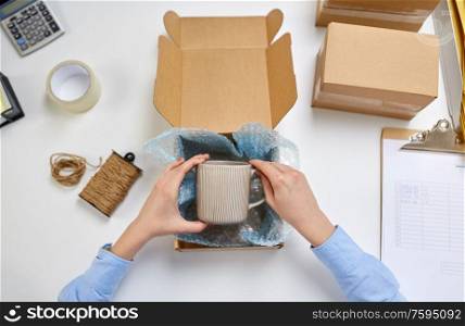 delivery, mail service, people and shipment concept - female hands packing mug into parcel box and it wrapping into protective bubble wrap at post office. hands packing mug to parcel box at post office
