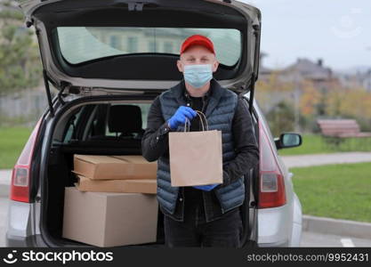 Delivery man in red cap, face medical mask and gloves hold paper bag near the car outdoor. Service coronavirus. Online shopping. mock up. Delivery man in red cap, face medical mask and gloves hold paper bag near the car outdoor. Service coronavirus. Online shopping. mock up.