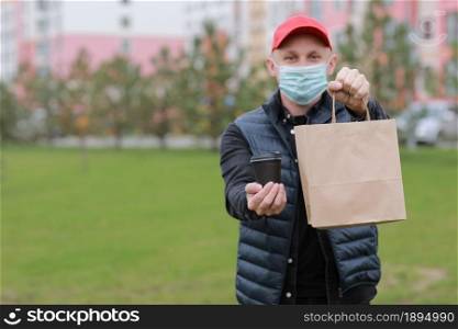 Delivery man in red cap, face medical mask hold take away paper bag and drink in disposable cup outdoors in city. Service coronavirus. Online shopping.. Delivery man in red cap, face medical mask hold take away paper bag and drink in disposable cup outdoors in city. Service coronavirus. Online shopping