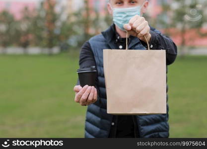 Delivery man in red cap, face medical mask hold take away paper bag and drink in disposable cup outdoors in city. Service coronavirus. Online shopping.. Delivery man in red cap, face medical mask hold take away paper bag and drink in disposable cup outdoors in city. Service coronavirus. Online shopping