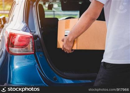 Delivery man is delivering cardboard box to customers via private car trunk door. People lifestyles and business occupation concept. Young male courier in casual clothes. Parcel move courier service