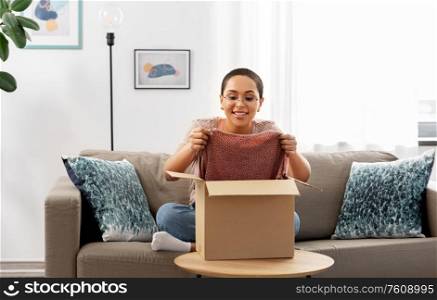 delivery, shipping and people concept - happy young african american woman opening parcel box with clothes at home. african american woman opening parcel box at home