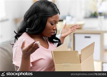 delivery, shipping and people concept - upset young african american woman opening parcel box with clothes at home. sad african american woman with parcel box at home