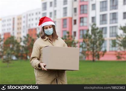 Delivery woman in a red Santa Claus hat and medical protective mask holds a big box outdoor. New Year&rsquo;s Christmas parcel in box, delivery online store in Quarantine time. Service coronavirus.. Delivery woman in a red Santa Claus hat and medical protective mask holds a big box outdoor. New Year&rsquo;s Christmas parcel in box, delivery online store in Quarantine time. Service coronavirus