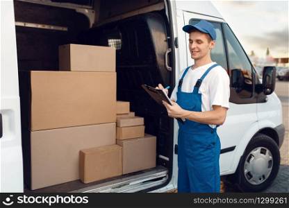 Deliveryman in uniform, carton boxes in the car, delivery service. Man standing at cardboard packages in vehicle, male deliver, courier or shipping job. Deliveryman in uniform, carton boxes in the car