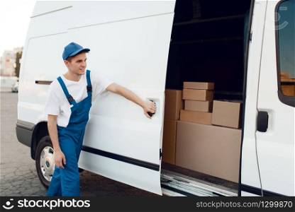 Deliveryman in uniform closing car door, auto with parcels and carton boxes, delivery service. Man standing at cardboard packages in vehicle, male deliver, courier or shipping job