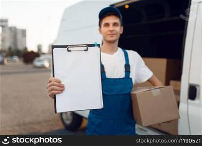 Deliveryman in uniform holding parcel and notebook, carton boxes in the car, delivery service. Man standing at cardboard packages in vehicle, male deliver, courier or shipping job