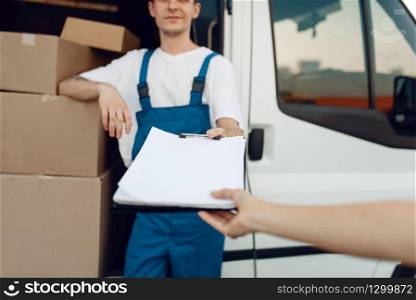 Deliveryman in uniform holding parcel and notebook, carton boxes in the car, delivery service. Man standing at cardboard packages in vehicle, male deliver. Deliveryman in uniform holding parcel, delivery