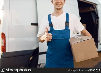 Deliveryman in uniform holding parcel and shows thumbs up, and notebook, carton boxes in the car, delivery. Man standing at cardboard packages in vehicle, male deliver, courier or shipping job. Deliveryman in uniform shows thumbs up, delivery