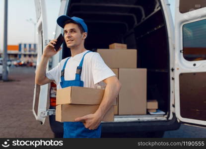 Deliveryman in uniform holds parcel and mobile phone at the car, delivery service, delivering. Man standing at cardboard packages in vehicle, male deliver, courier or shipping job