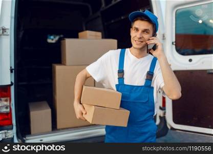 Deliveryman in uniform holds parcel and mobile phone at the car, delivery service, delivering. Man standing at cardboard packages in vehicle, male deliver, courier or shipping job. Deliveryman in uniform holds parcel and phone