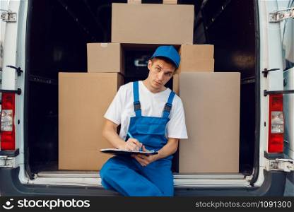 Deliveryman in uniform, parcels in the car on background, cargo, delivery service. Man standing at cardboard packages in vehicle, male deliver, courier or shipping job. Deliveryman in uniform, parcels in the car, cargo
