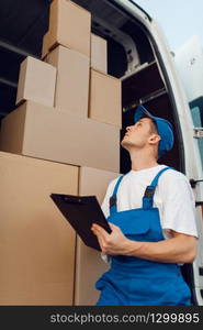 Deliveryman in uniform, parcels in the car on background, cargo, delivery service. Man standing at cardboard packages in vehicle, male deliver. Deliveryman in uniform, parcels in the car, cargo