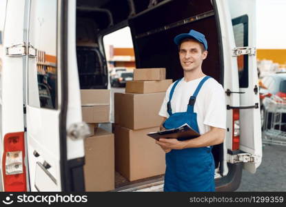 Deliveryman in uniform poses at the car with parcel boxes, delivery service. Man standing at cardboard packages in vehicle, male deliver, courier or shipping job