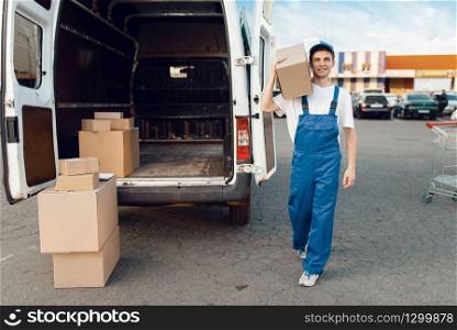 Deliveryman in uniform poses at the car with parcel boxes, delivery service. Man standing at cardboard packages in vehicle, male deliver, courier or shipping job. Deliveryman poses at the car with parcel boxes