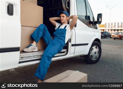 Deliveryman in uniform relaxing in the car during a break, auto with parcels and carton boxes, delivery service. Man poses at cardboard packages in vehicle, male deliver, courier or shipping job. Deliveryman relaxing in the car during a break