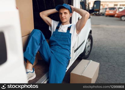 Deliveryman in uniform relaxing in the car during a break, auto with parcels and carton boxes, delivery service. Man poses at cardboard packages in vehicle, male deliver, courier or shipping job. Deliveryman relaxing in the car during a break