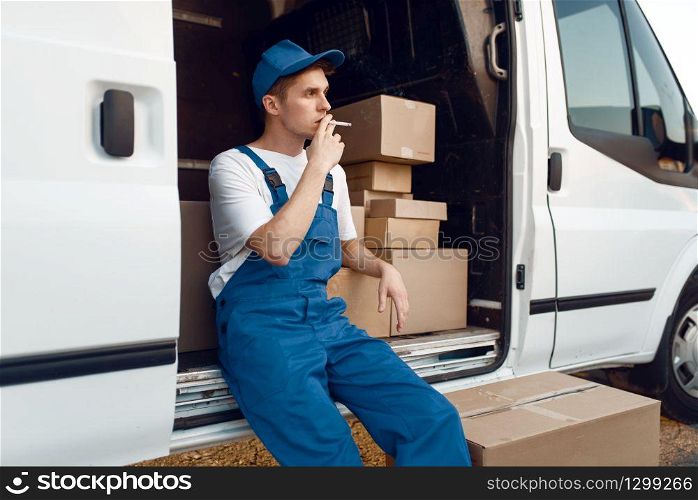 Deliveryman in uniform smokes at the car during a break, auto with parcels and carton boxes, delivery service. Man standing at cardboard packages in vehicle, male deliver, courier or shipping job