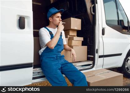 Deliveryman in uniform smokes at the car during a break, auto with parcels and carton boxes, delivery service. Man standing at cardboard packages in vehicle, male deliver, courier or shipping job
