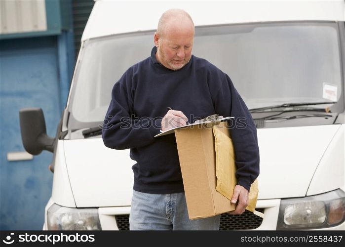 Deliveryperson standing with van writing in clipboard holding box smiling