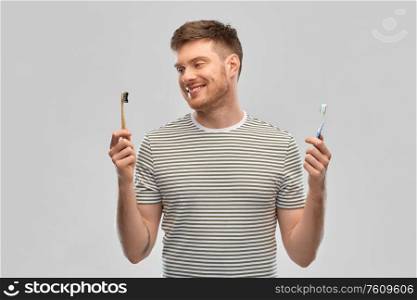 dental care, oral hygiene and people concept - happy smiling young man comparing wooden and plastic toothbrush over gray background. man comparing wooden and plastic toothbrush