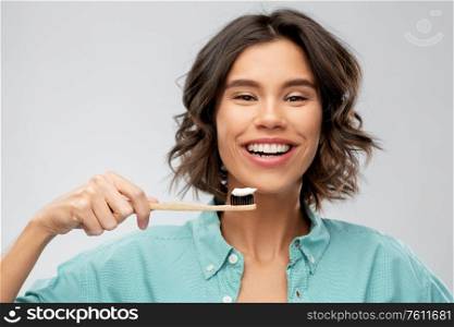 dental care, oral hygiene and people concept - portrait of happy smiling young woman in turquoise shirt with toothpaste on wooden toothbrush over gray background. smiling woman with toothpaste on wooden toothbrush