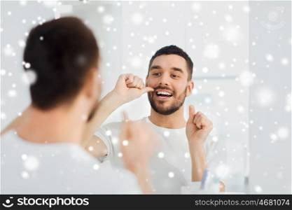 dental care, people, winter, christmas and hygiene concept - smiling young man with floss cleaning teeth and looking to mirror at home bathroom over snow