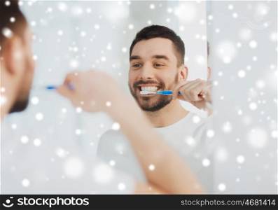 dental care , winter, christmas people and hygiene concept - smiling young man with toothbrush cleaning teeth and looking to mirror at home bathroom over snow