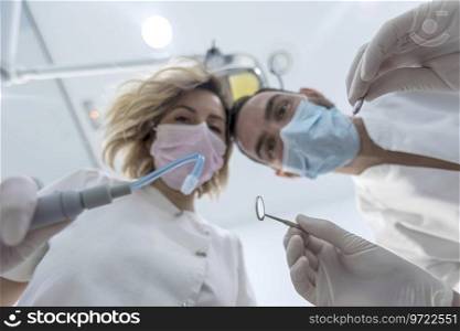 Dentist and his nurse leaned above the patient. Doctor in mask applies metal instruments. Low angle view from patient’s perspective.
