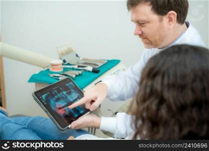 Dentist are showing X-ray on a screen to his patient.
