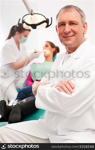 Dentist in his surgery looking at the viewer, in the background his assistant is giving a female patient a treatment
