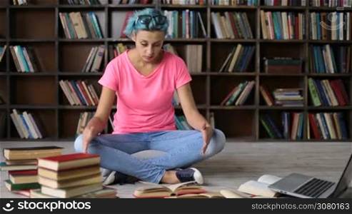 Depressed college female student with blue hair surrounded by books sits on the floor cross legged. Frustrated hipster girl with eyeglasses on her head studying complicated material in university library and preparing for exam.