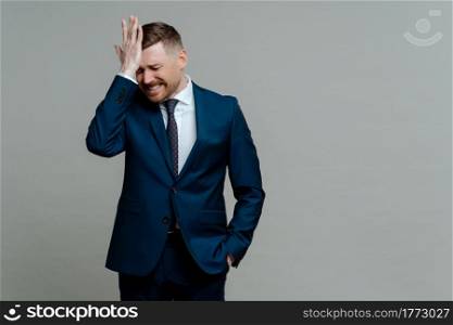 Depressed desperate businessman in suit getting bad news, touching forehead with hand and feeling upset worried while standing against grey background. Ceo executive manager having problems at work. Depressed businessman in suit receiving bad news and feeling worried