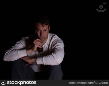 Depressed man holding bottle of beer against face while in thought. Dark background.