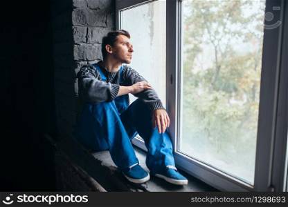 Depressed man sitting on the window sill in dark room, psycho patient. Mentally ill people concept, stressed human, depression