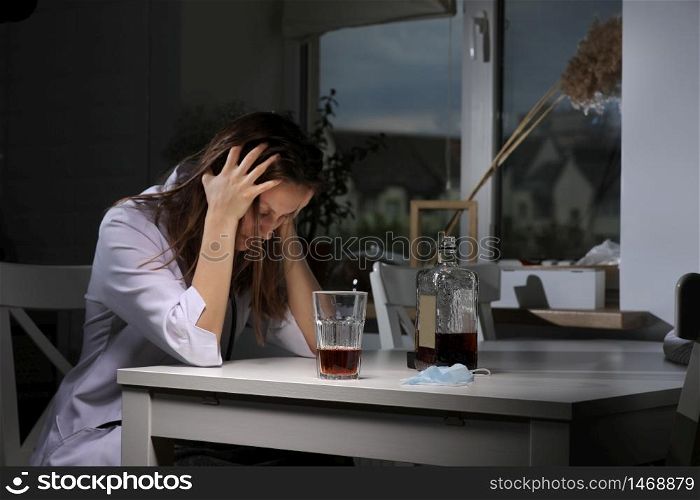 depressed tired female doctor sits at the table at home holds hands behind head and drinks strong alcohol after working in hospital during coronavirus.. depressed tired female doctor sits at the table at home holds hands behind head and drinks strong alcohol after working in hospital during coronavirus