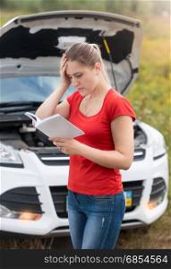 Depressed woman looking at her broken car and reading car owner manual