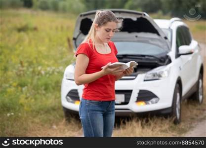 Depressed woman looking at her broken car and reading owner manual