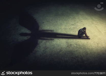 Depressed young man sitting alone isolated in a dark room, casting a shadow of a powerful guardian angel. Inner strength conceptual background. Stressed person introvert with an imaginary friend