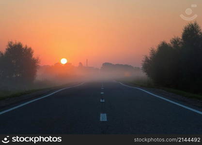 Deserted straight asphalt road disappears into the morning mist at dawn. The golden disk of the sun rises in a cloudless orange sky. Sunrise in the early foggy morning.