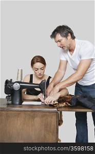 Designer helping coworker in stitching cloth on sewing machine over colored background